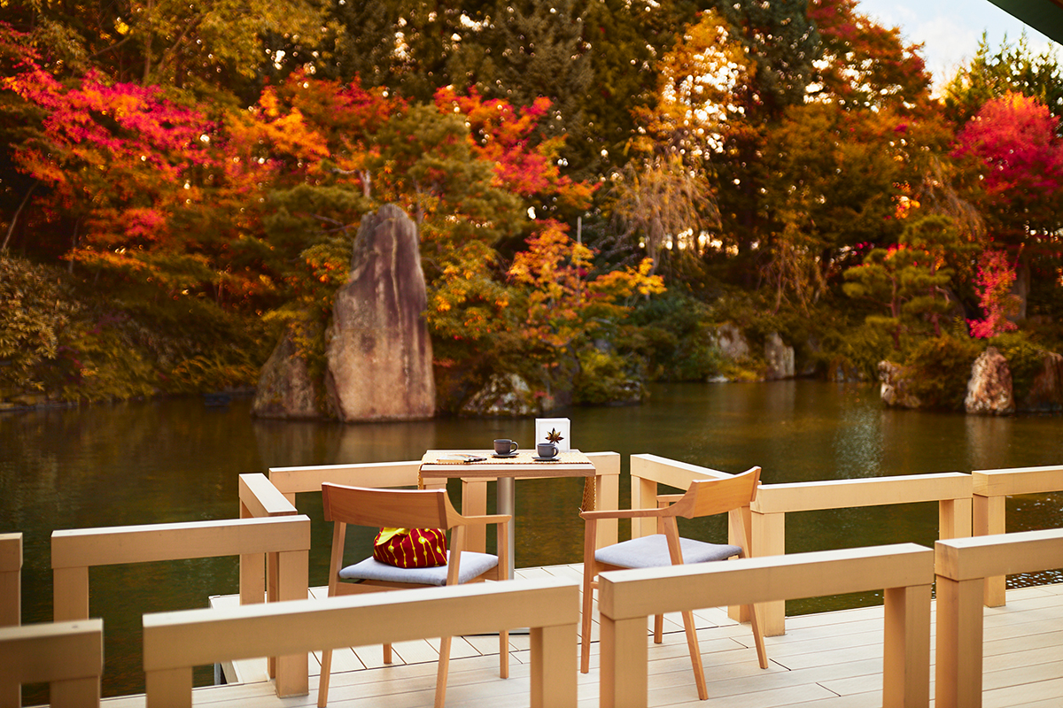 手前の通路に椅子とテーブルが置かれ、その向こうに水面、そのさらに奥に紅葉した木々が広がる昼間の水庭の写真