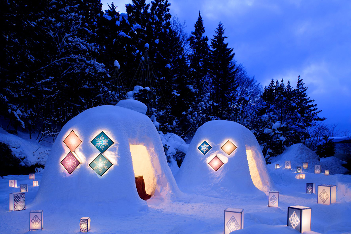 雪が積もった景色の中にかまくらがあり、こぎん燈籠でライトアップされた夕暮れの水庭の写真