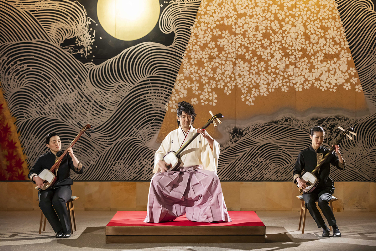 壁画「春秋波濤」を背景に、真ん中に渋谷幸平氏、その左右に女性が座って三味線を演奏している写真