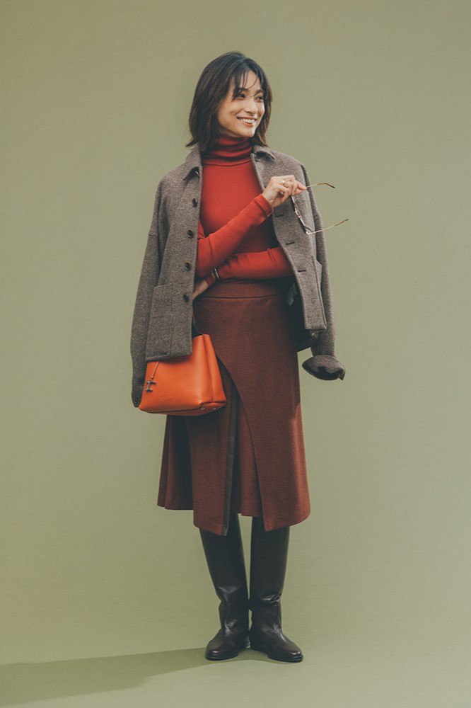 明るい赤茶のタートルニットと暗めの赤茶のスカートを着て、肩に薄茶色のコートをかけ、ロングブーツを履いたモデル画像