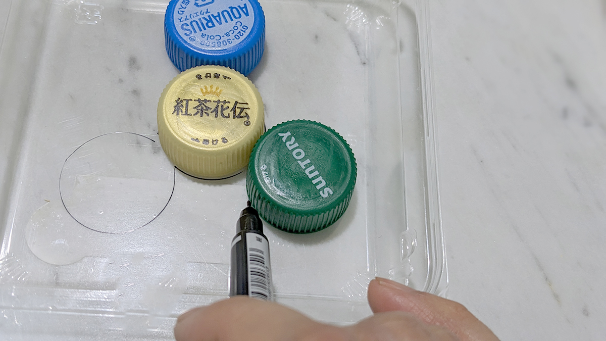 プラ容器の蓋の上にペットボトルのキャップを並べ、油性ペンで輪郭をなぞっている写真　中心にひとつのキャップ、その周囲3箇所にキャップを等間隔に並べる