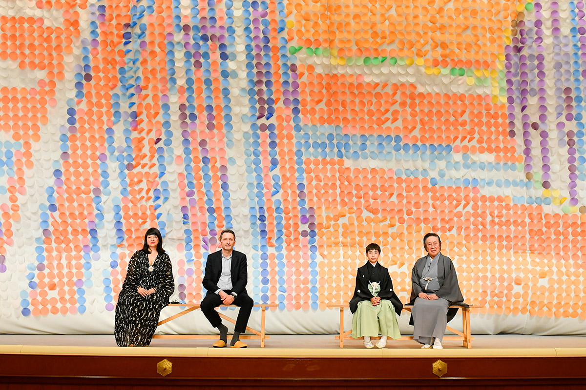 オレンジやブルーなどの色の丸いパーツが敷き詰められた祝幕を背景に座る4人の写真