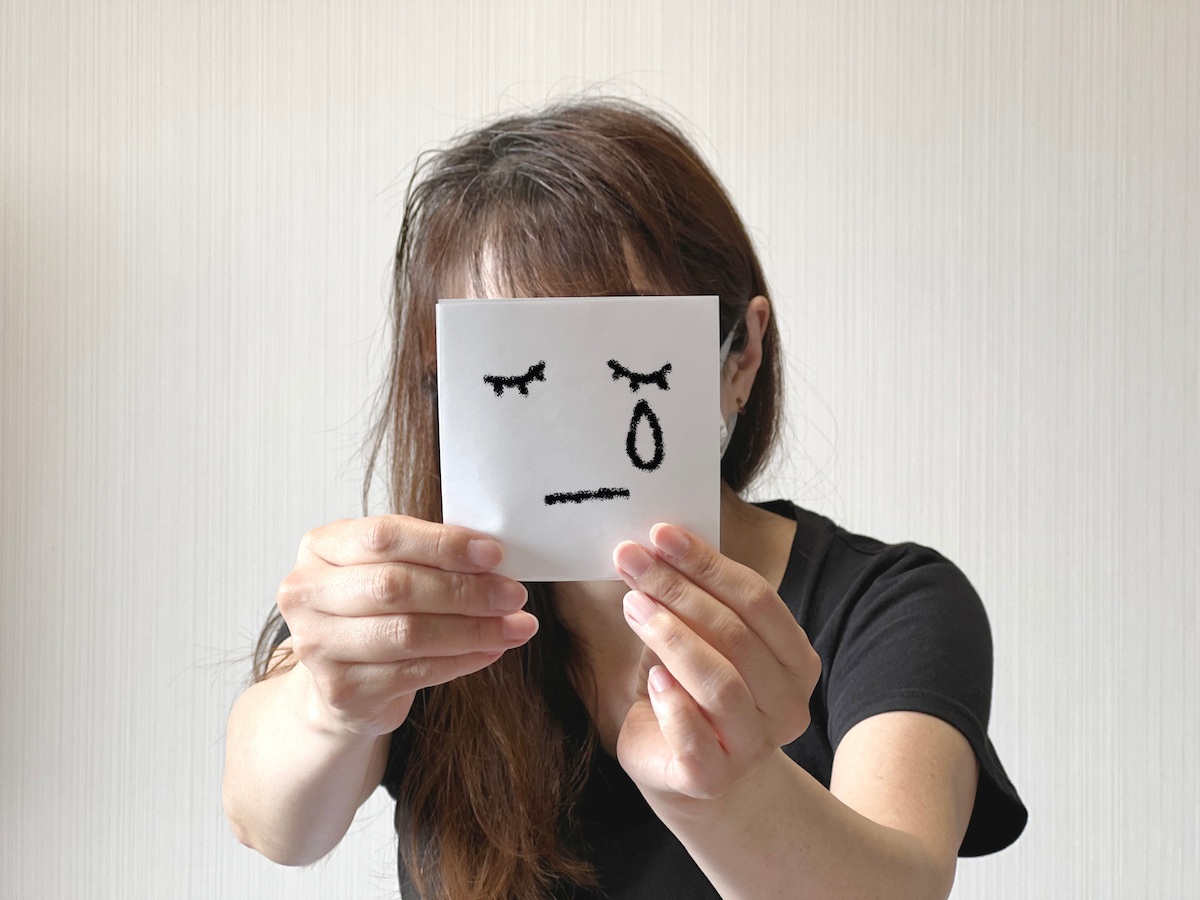 泣いている顔のイラストが描かれた紙を顔の前にかざしている人の写真