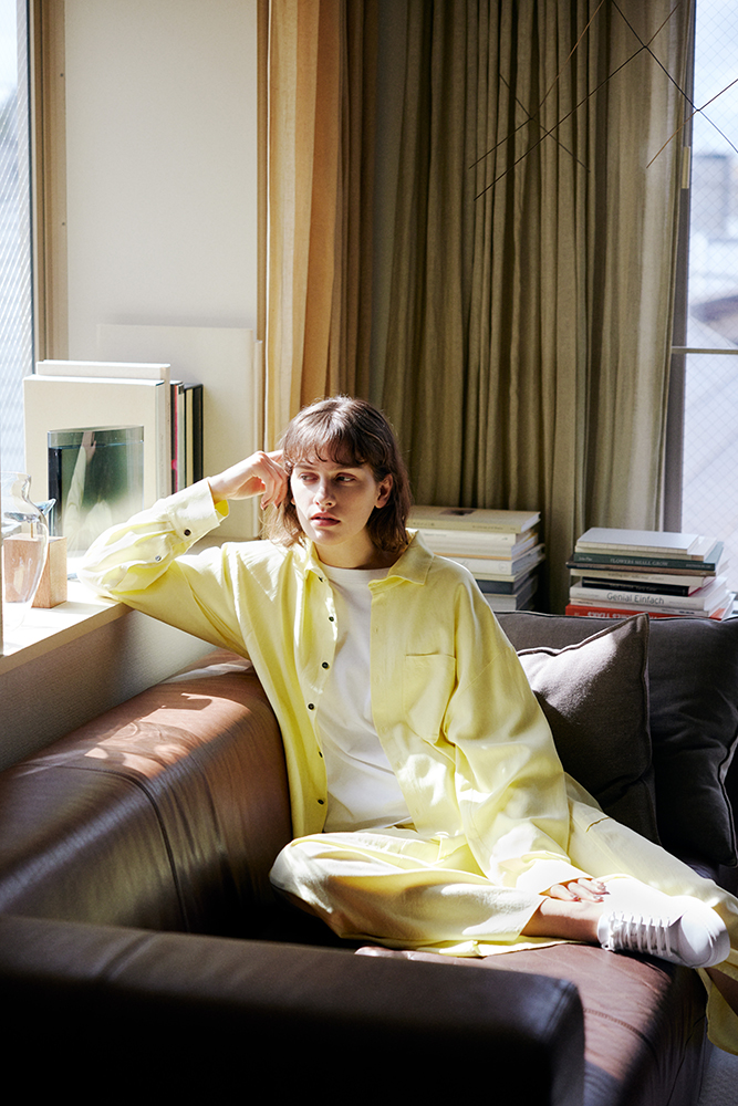 白いトップスに黄色のシャツを羽織り、ソファに座っているモデル写真