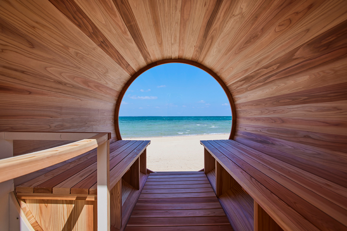 丸い窓の向こうに海と砂浜が見えるサウナの中の写真