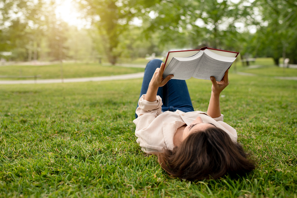 芝生の上に寝転んで本を広げている人の写真