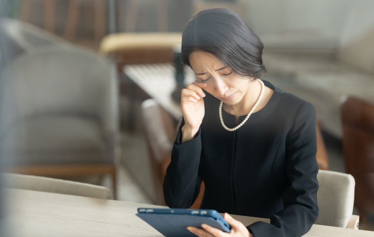 個人との思い出をタブレットで振り返りながら涙する喪服姿の女性の写真