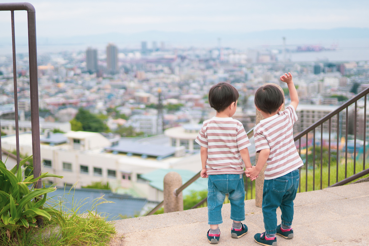 山の上の公園の展望台の階段上から街並みを見ながら手を振っている日本の双子兄弟。奥に海が広がっている。薄い雲がかかった天気