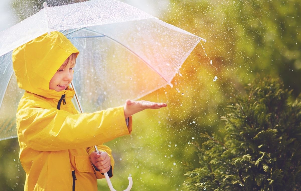 黄色の合羽を着て右手に傘を持ち、左手で雨を受け止める外国人の男の子の写真