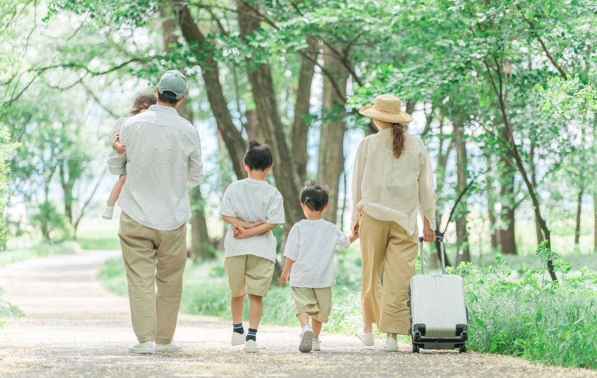 スーツケースを持った4人家族が広い道を並んで歩いている後姿の写真