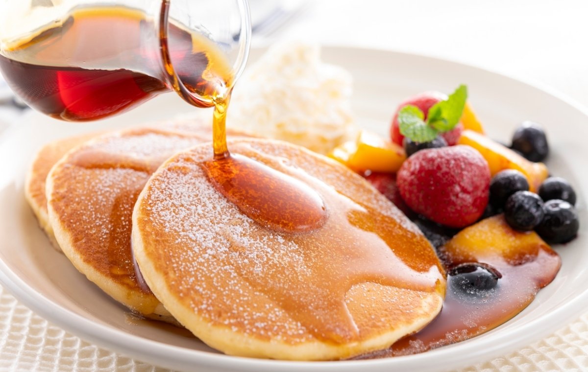 フルーツ・生クリームが添えられたパンケーキにメープルシロップをかけている写真