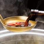 湯気が立つ鍋の上で、醤油瓶からおたまに醬油を注いでいる写真