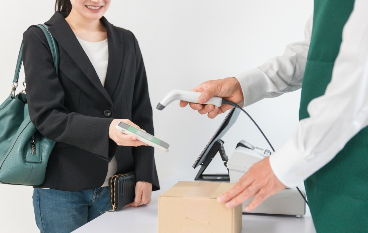 コンビニのレジで荷物を受け取るため、スマホのバーコードをスキャンしてもらう女性の写真